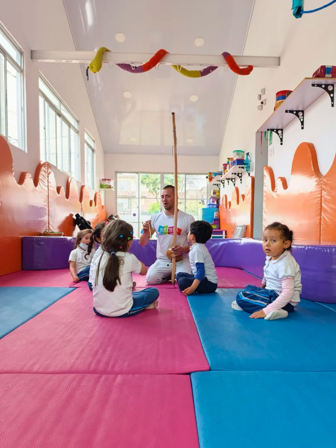 Clase de música y capoeira: instructor con berimbau y niños sentados en colchonetas coloridas